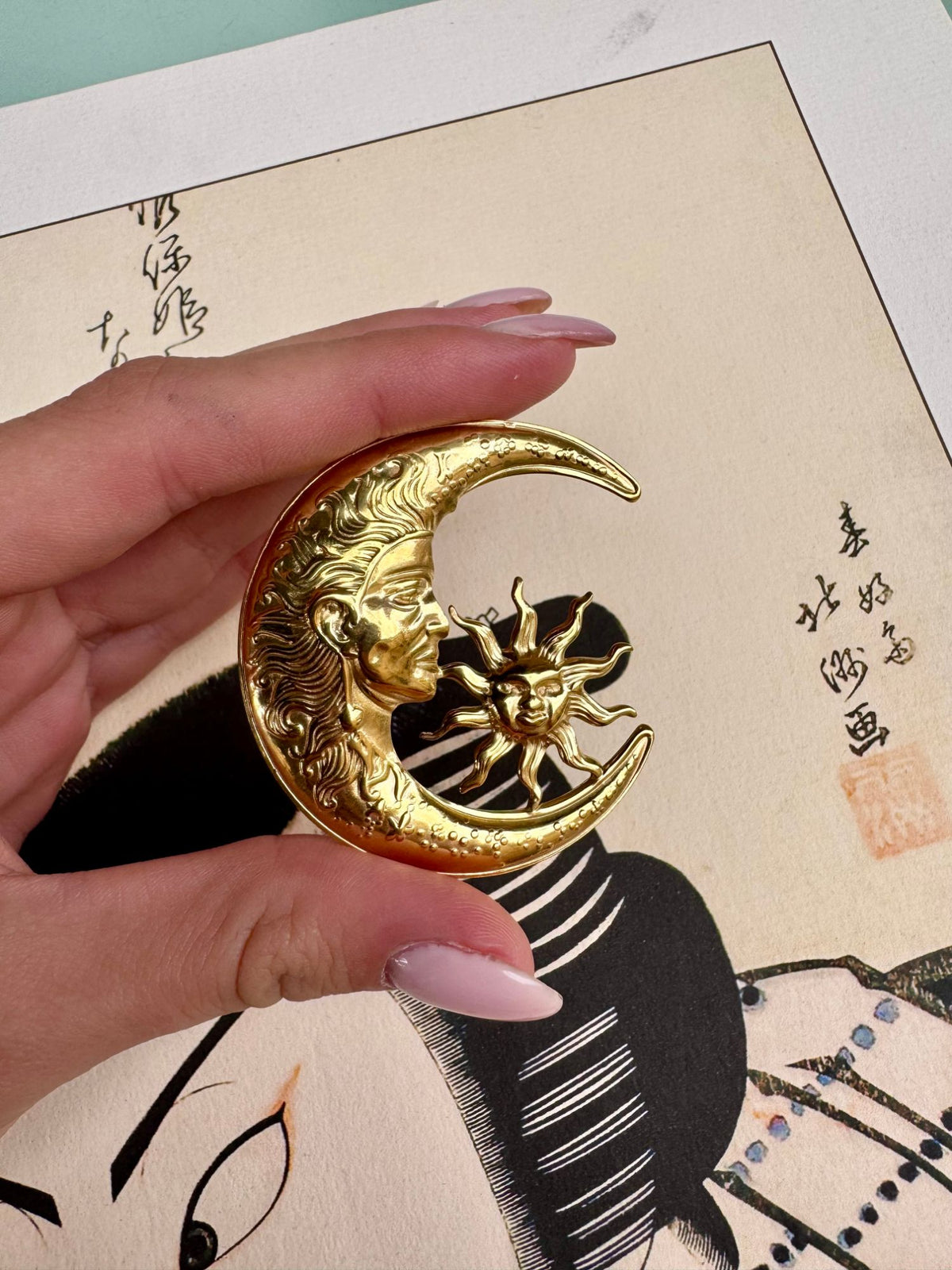 Broche lune et soleil dorée tenue en main, détail du visage de la lune et du soleil en relief